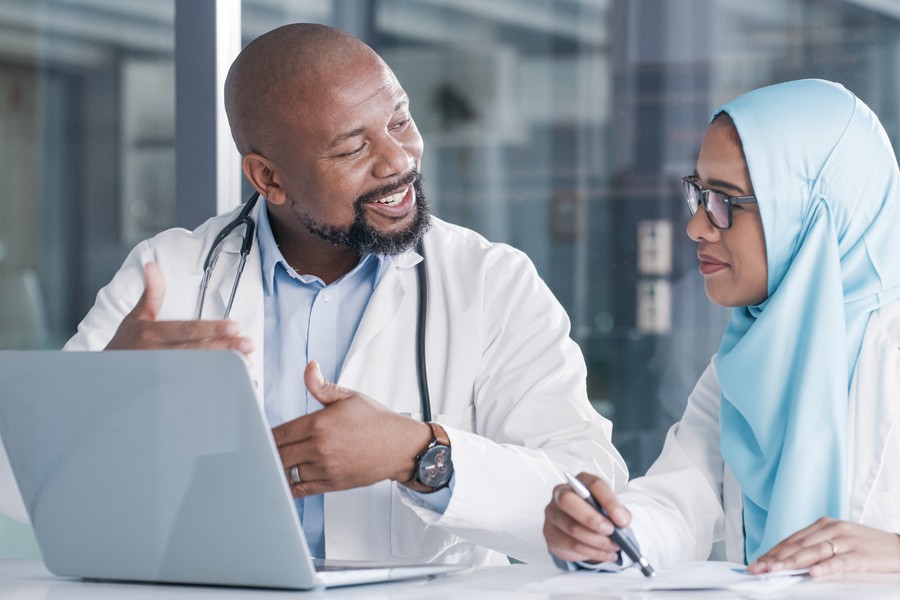 A male doctor talks to a female colleague wearing a hijab, both engaged in conversation by a laptop.
