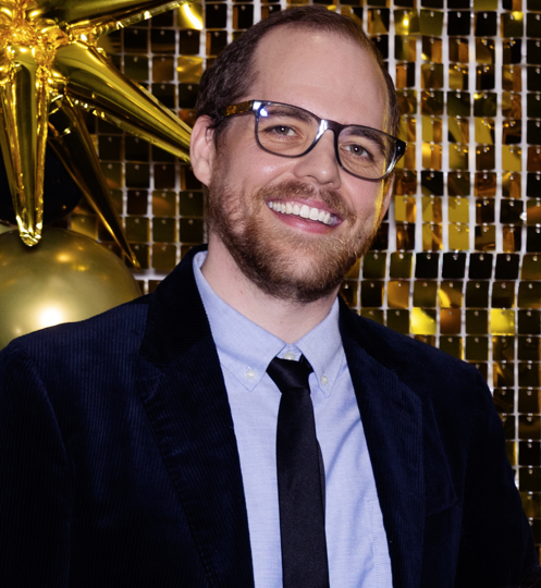Smiling man with glasses in navy blazer and tie before gold balloons and sequin backdrop