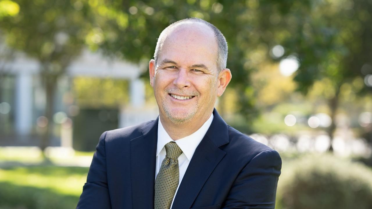 Jim Hankins smiling at camera, wearing suit and tie, photo taken outdoors with trees in background.