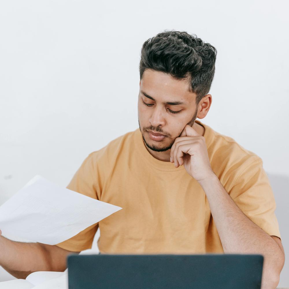 A man sits in front of a laptop. He is holding documents and is thinking deeply.