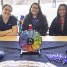 Three smiling women sit behind a table with a colorful prize wheel and promotional items.