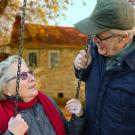 An older man smiles down at an older woman while she sits on a swing.