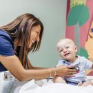 A smiling female nurse listens to a baby's heart with a stethoscope.